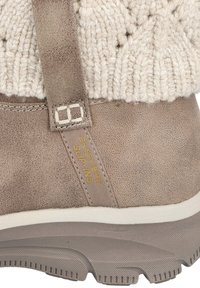 Acercamiento a una bota de invierno taupe de tipo slip-on con puño de tejido y suela de goma, con la etiqueta "Skechers Slip-Ins" en el lado.