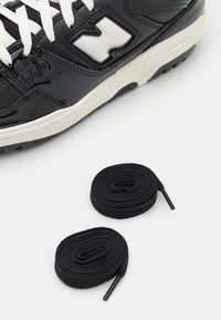Sneaker in pelle nera con finitura lucida, lacci bianchi, grande logo e suola in gomma grigia. Due lacci rotondi neri accanto alla scarpa.