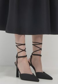 Donna che indossa scarpe con tacco a punta nere con cinturini alla caviglia, abbinate a una gonna midi nera su uno sfondo semplice.