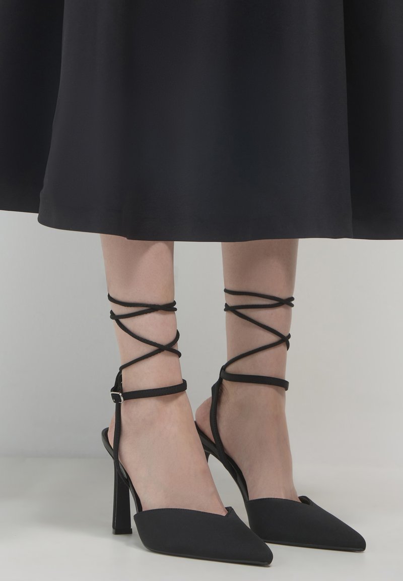 Donna che indossa scarpe con tacco a punta nere con cinturini alla caviglia, abbinate a una gonna midi nera su uno sfondo semplice.