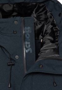 Donkerblauw marinejack met rits, voorzien van een merklabel aan de binnenkant, trekkoord en glanzende zwarte voering. Getextureerd buitenmateriaal, functionele hardware.