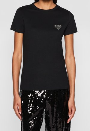 Vrouw draagt een zwarte aansluitende t-shirt met een klein zilveren hartembleem op de linkerborst, gecombineerd met zwarte paillettenbroek.