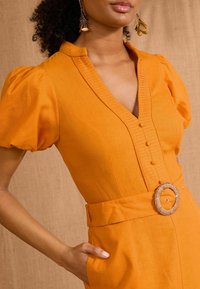 Rochie din in portocalie cu mâneci bufante, decolteu în V adânc, nasturi în față și o curea cu o cataramă rotundă țesută. Material texturat vizibil.