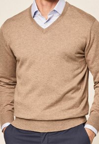 Beige v-neck sweater made of soft fabric with long sleeves, ribbed cuffs, and hem, paired with a light blue collared shirt underneath.