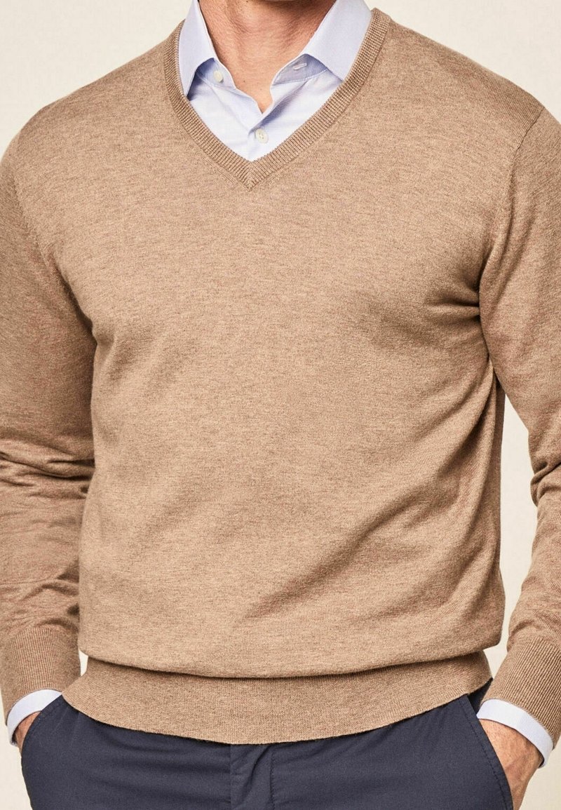 Beige v-neck sweater made of soft fabric with long sleeves, ribbed cuffs, and hem, paired with a light blue collared shirt underneath.