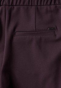 Tissu violet foncé avec une texture lisse. Présente une taille élastique et une petite poche arrière rectangulaire avec une étiquette de marque.