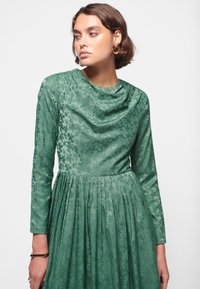 Grønne blomstermønstrede kjole med lange ærmer, draperet halsudskæring og plisseret skørt. Stoffet har en glat tekstur og en subtil glans.