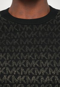 Maglione nero a maglia con un motivo testurizzato di loghi "KM" intrecciati in oro metallico. Il colletto a costine aggiunge un tocco di dettaglio.