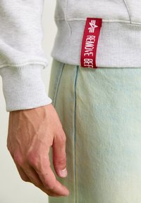 Szürke pulóver bordás mandzsettával, fehér szöveggel ellátott piros címkével. Világoskék farmernadrág látható, sima textúrával és szűkített kivágással.
