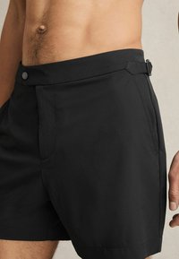 Egy férfi törzsének közeli képe, fekete, szabott rövidnadrágot visel, amely gombbal, oldalsó állítóval és elülső zsebekkel rendelkezik, az oldalán bőr és kéz látható.