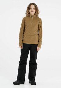 Smeđi pull-over od flisa s polu-zip ovratnikom i logotipom, u kombinaciji s crnim hlačama i čvrstim crnim čizmama. Model stoji ispred bijelog pozadinskog zida.