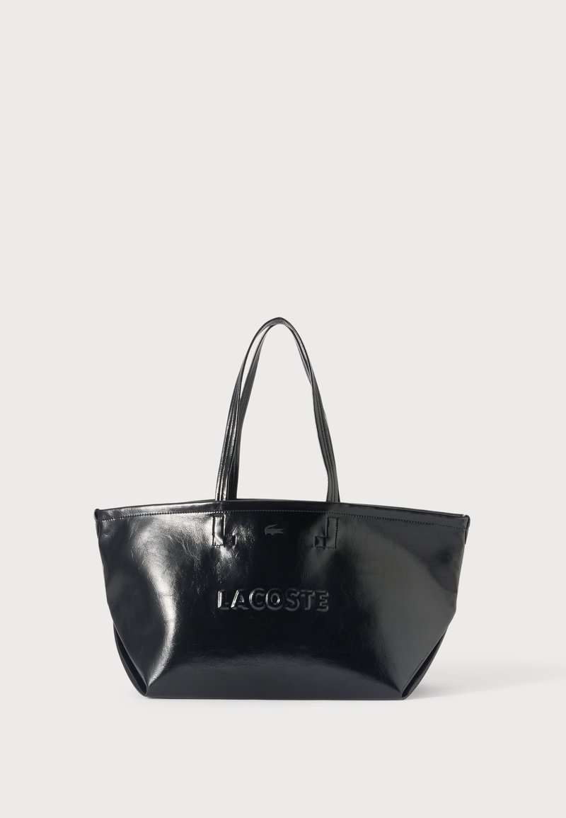 Λαμπερή μαύρη τσάντα tote Lacoste με δύο λαβές και ανάγλυφο όνομα επωνυμίας στο μπροστινό μέρος, εμφανιζόμενη σε απλό ανοιχτό φόντο.