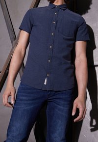 Kortärmad, marinblå skjorta med ett strukturerat mönster, knappstängning och bröstficka. Matchas med mörka jeans.