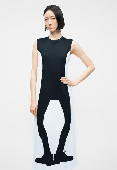 Γυναίκα που φοράει αμάνικο ασπρόμαυρο φόρεμα με εκτυπωμένη μαύρη σιλουέτα ποδιών και παπουτσιών μπροστά.