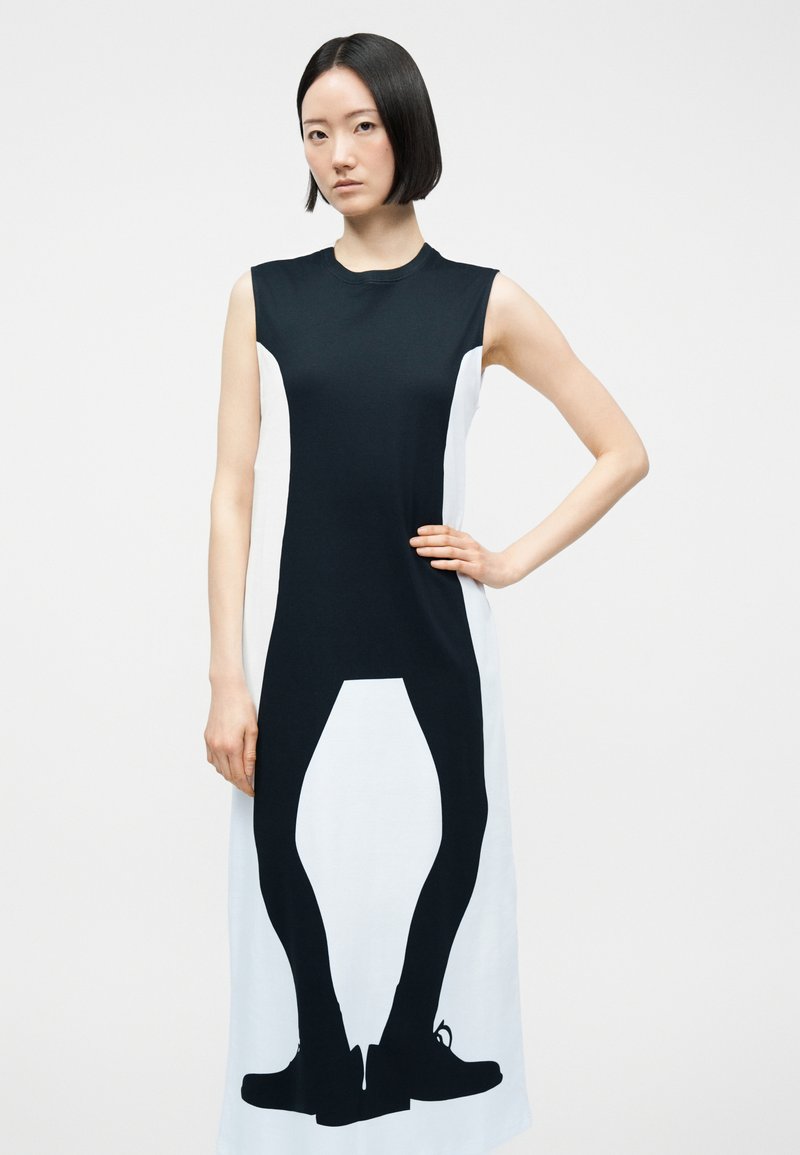 Femme portant une robe sans manches noire et blanche avec une silhouette imprimée en noir représentant des jambes et des chaussures à l'avant.