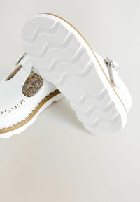 Sandales en cuir blanc avec détail floral, fermeture à boucle, et semelle en caoutchouc texturé présentant un motif ondulé et une finition contrastante.