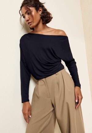 Kvinde iført en sort off-shoulder langærmet top og beige højtaljede vide bukser mod en lys væg baggrund.