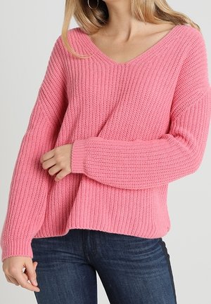 Donna che indossa un maglione a costine rosa con scollo a V e maniche lunghe, pantaloni jeans blu scuro, in posa con un braccio incrociato e l'altro rilassato.