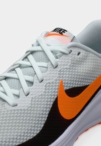 Chaussure de course Nike gris clair avec une tige en mesh, des accents texturés, un logo Swoosh orange, des lacets plats et une semelle blanche amortie.