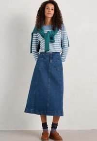 Pruhované modro-bílé tričko s dlouhým rukávem, s zeleným svetrem uvázaným na krku, v kombinaci s modrou džínovou sukní až na zem a hnědými botami.