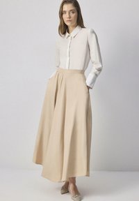 Woman wearing a white long-sleeve blouse tucked into a high-waisted beige asymmetrical midi skirt and beige pointed flats with embellishments.