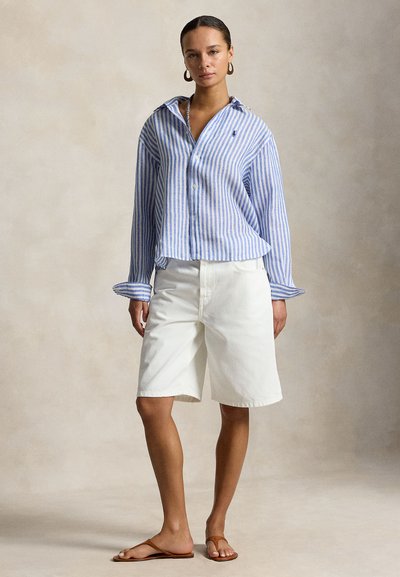 Nő kék-fehér csíkos gombos ingben, fehér térdig érő rövidnadrágban, barna szandálban és karika fülbevalóval bézs háttér előtt áll.