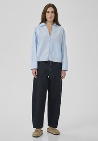 Kvinna som står iklädd en ljusblå oversize-skjorta, mörkblå vida jeans och beiga slip-on-skor mot en enkel ljusgrå bakgrund.