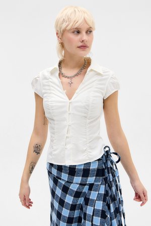 Lühikeste blondide juustega naine kannab valget keha järgivat nööbitavat pluusi, hõbedast ketti risti kujutisega ja sinakas-mustruudilist volditud seelikut.