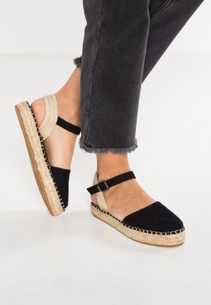 Espadrilles de lienzo negras con suela de yute, correa en el tobillo y detalle tejido en contraste a lo largo del borde. Llevadas con jeans negros cortos.