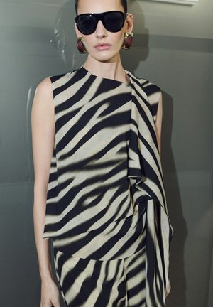 Femme portant un haut sans manches et un pantalon avec des rayures zébrées noires et blanches, de grandes boucles d'oreilles dorées et bordeaux, et des lunettes de soleil noires oversize.