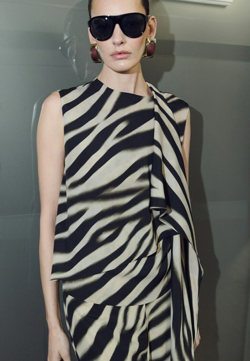 Femme portant un haut sans manches et un pantalon avec des rayures zébrées noires et blanches, de grandes boucles d'oreilles dorées et bordeaux, et des lunettes de soleil noires oversize.