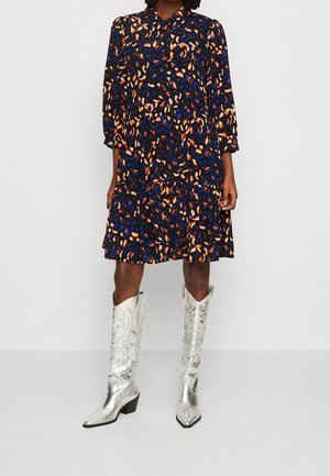 Kvinde iført en mørkeblå kjole med orange og pink abstrakt print og sølvfarvede cowboy-støvler til knæet, stående mod en ensfarvet hvid baggrund.
