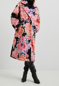 Kabát s květinovým vzorem v růžových a černých odstínech, dvojitý řádek knoflíků, velké klopy a volný střih, doplněný černými kozačkami sahajícími ke kolenům.