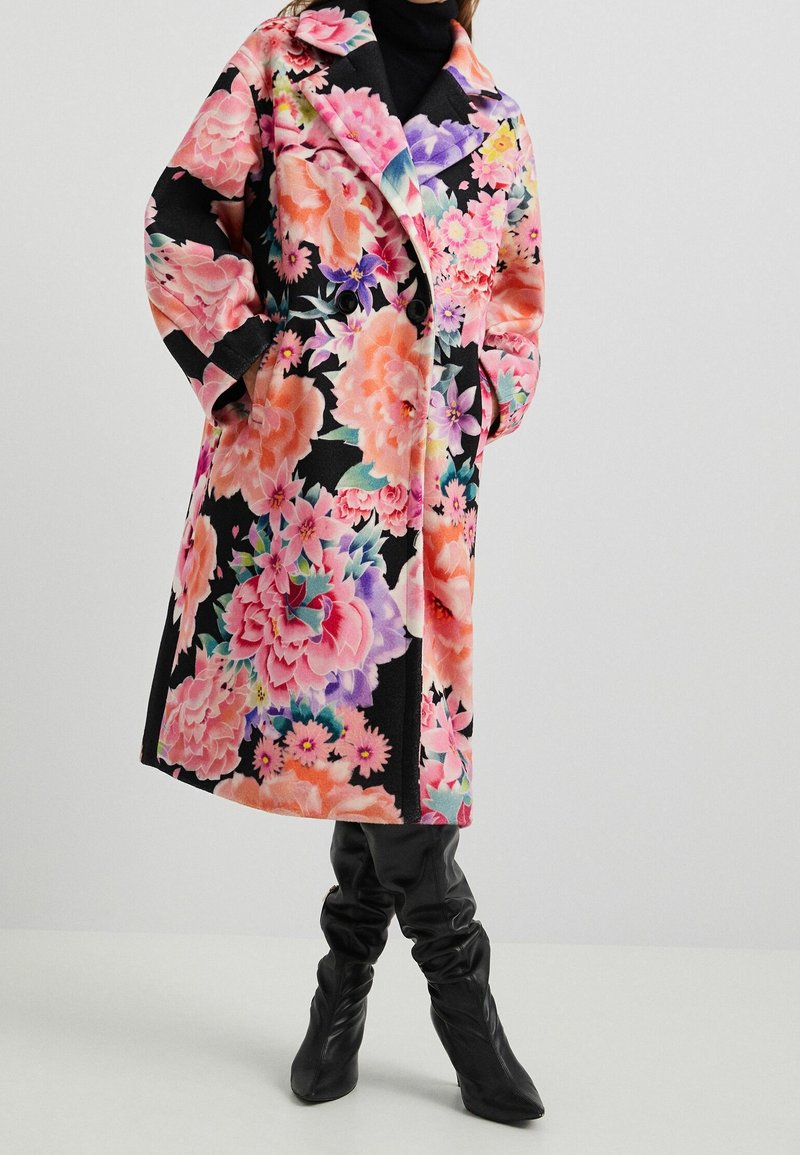 Kabát s květinovým vzorem v růžových a černých odstínech, dvojitý řádek knoflíků, velké klopy a volný střih, doplněný černými kozačkami sahajícími ke kolenům.