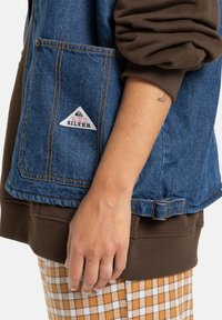Personens hånd ved siden av en blå denim Quiksilver vest over en brun erme og rutete gul-hvite bukser.