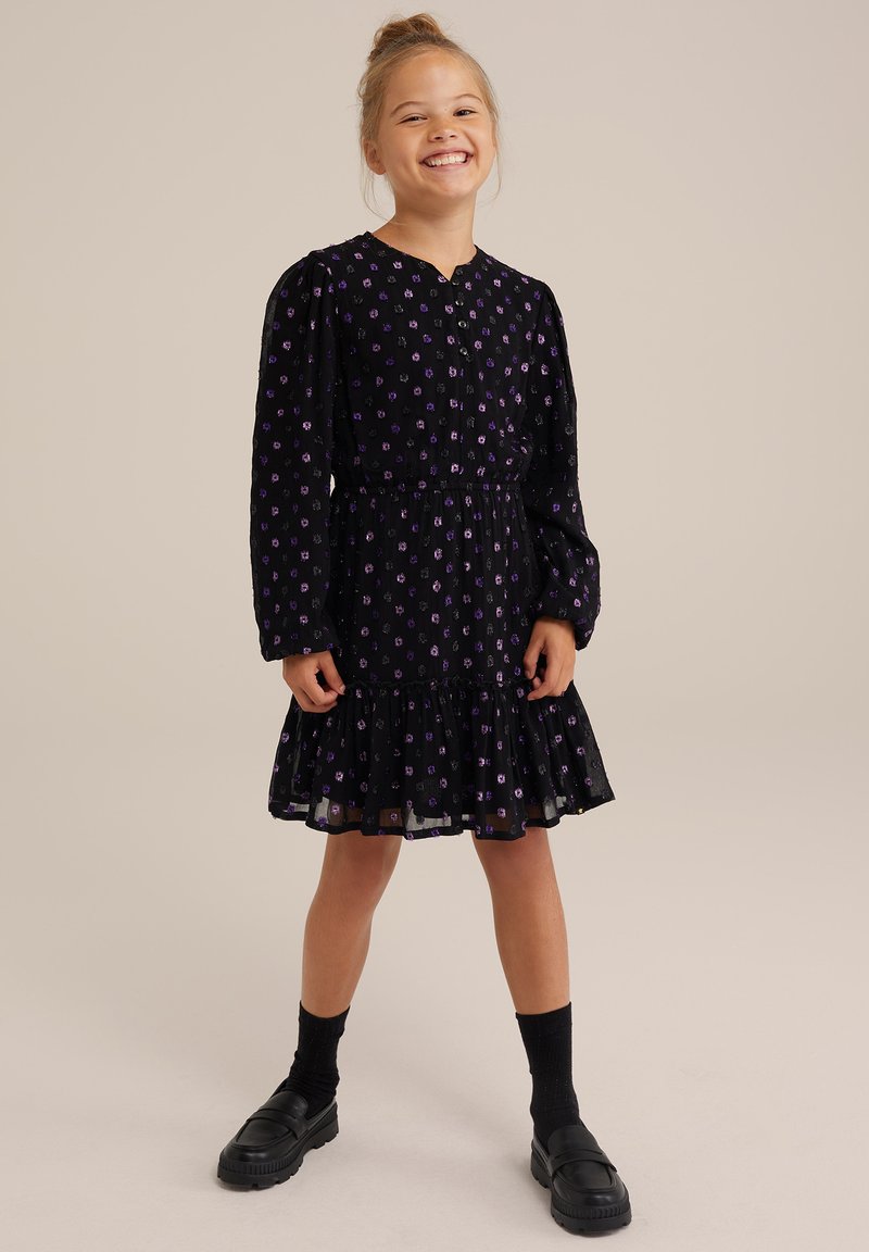 Schwarzes Kleid mit lila Blumenmuster, langen Ärmeln, Rüschensaum, tailliertem Schnitt und Knopfleiste. Getragen mit schwarzen Plateauschuhen und Socken.