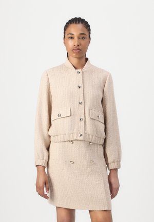 Veste en tweed beige avec un col, des boutons sur le devant et deux poches. Assortie à une jupe texturée correspondante dotée de boutons à double boutonnage.