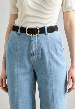 Cintura nera con fibbia a doppio anello metallico, indossata sopra jeans in denim di un blu chiaro a vita alta con gamba dritta. Tessuto texturizzato.