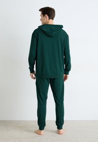 Mann som står barbeint på teppet med ryggen til, iført mørkegrønn hettegenser og matchende joggebukser foran en plain hvit vegg.