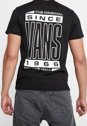 Homme portant un t-shirt Vans noir avec un grand texte blanc « The Original Since 1966 Vans Off The Wall » au dos et un jean gris foncé.