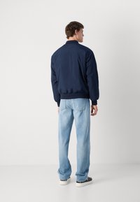 Veste bomber bleu marine avec une texture lisse, des poignets et un col côtelés. Un jean droit bleu clair et des baskets noires complètent la tenue.