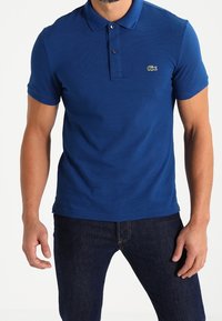 Blue polo shirt with a textured finish, short sleeves, and a two-button placket. Features a small green Lacoste logo on the chest.