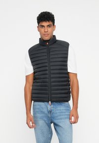 Sort polstret vest med høj krave, lynlås foran, med vandrette kanaler og en orange lynlås træk. Båret over en hvid t-shirt med blå jeans.