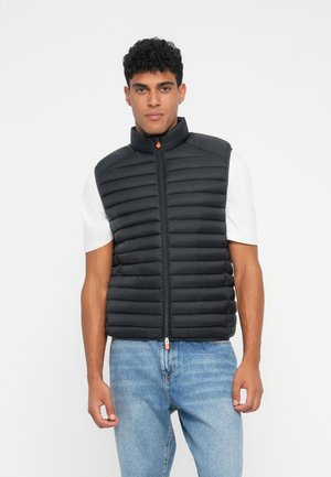 Zwarte gewatteerde vest met hoge kraag, rits aan de voorkant, met horizontale stiksels en een oranje ritslip. G gedragen over een wit T-shirt met blauwe jeans.