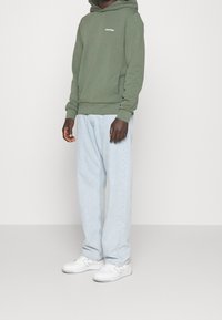 Zelená bavlněná mikina s potiskem na přední straně, kombinovaná se světle modrými džíny a bílými teniskami. Jednoduchý design, volný střih a měkká textura.