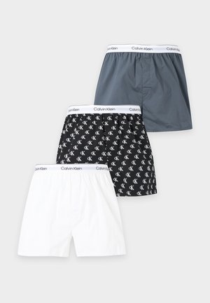 Homme mince à la peau foncée debout pieds nus, portant un boxer gris Calvin Klein, regardant légèrement vers sa gauche sur un fond clair uni.