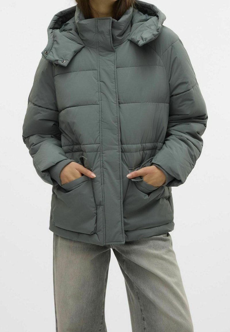 Veste matelassée grise avec capuche, présentant une texture matelassée, une fermeture éclair à l'avant et des poches latérales, portée avec un pantalon gris clair.