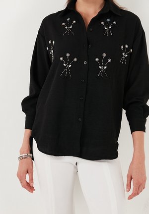 Femme portant une chemise noire à boutons avec de petits motifs floraux brodés et un pantalon blanc, montrée du cou jusqu'aux hanches.