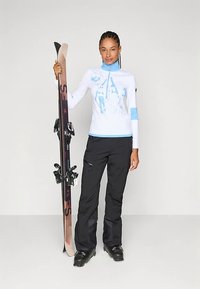 Síruha, amely fehér, hosszú ujjú felsőből áll kék díszítéssel és grafikával, fekete sínadrággal és fekete síbakancsokkal, síléceket tartva.