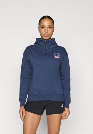 Felpa blu navy con zip e collo alto, con logo rosso e bianco sul petto e polsini e orlo a coste. Indossata con pantaloncini neri.